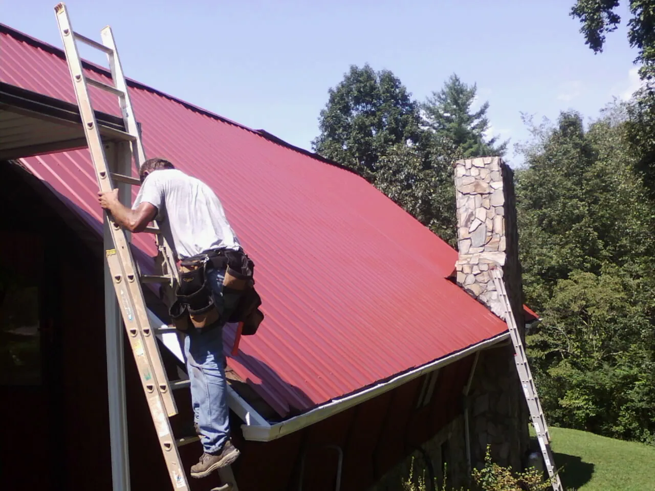 Roofer on a ladder beside a red metal roof and stone chimney — Soffit & Fascia Repair by Roofing Brothers in Cutchogue